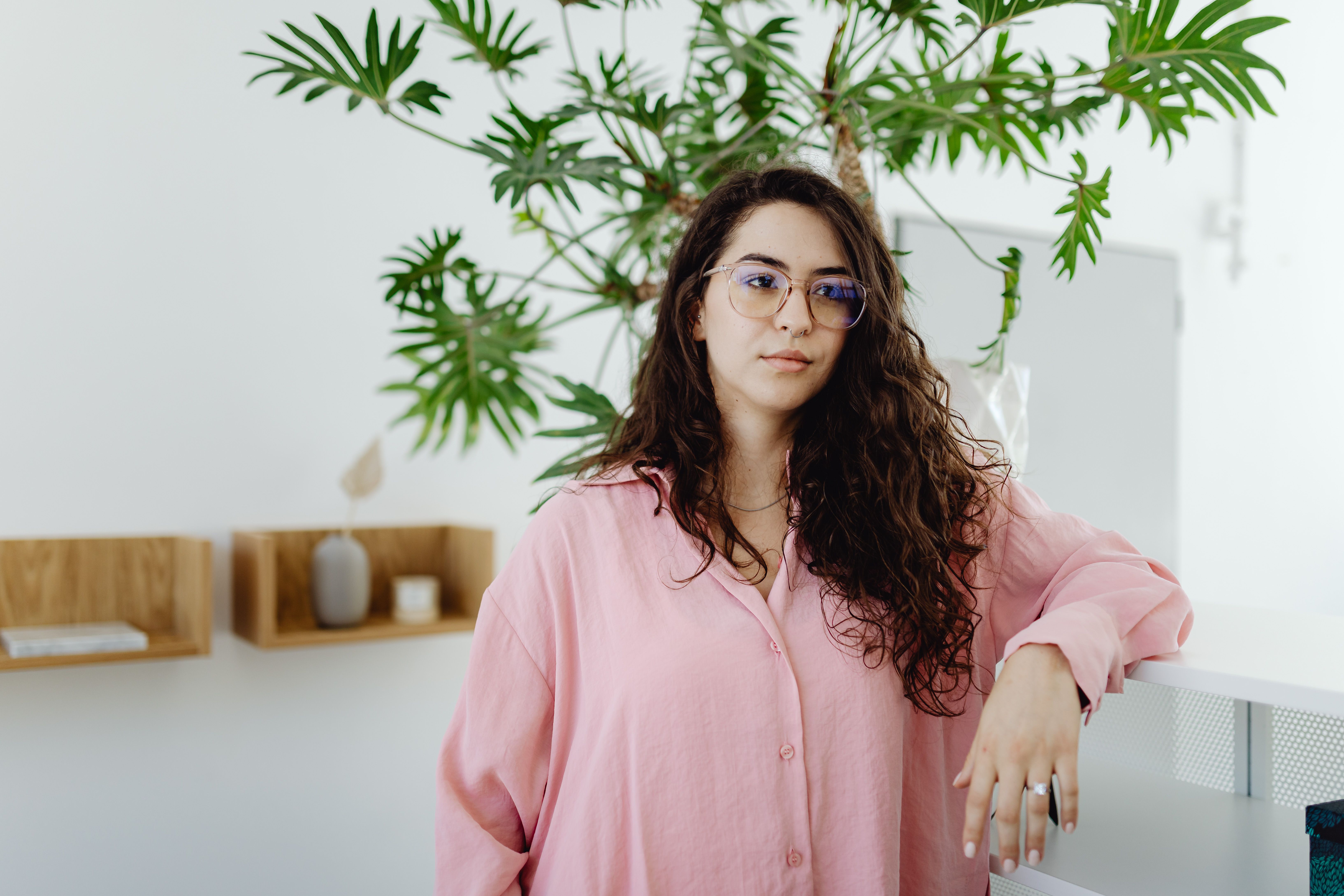 A woman with curly hair and eyeglasses standing confidently in a modern office space with plants.