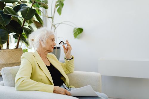 Free Elderly businesswoman seated with papers and glasses, indoors with plants. Stock Photo
