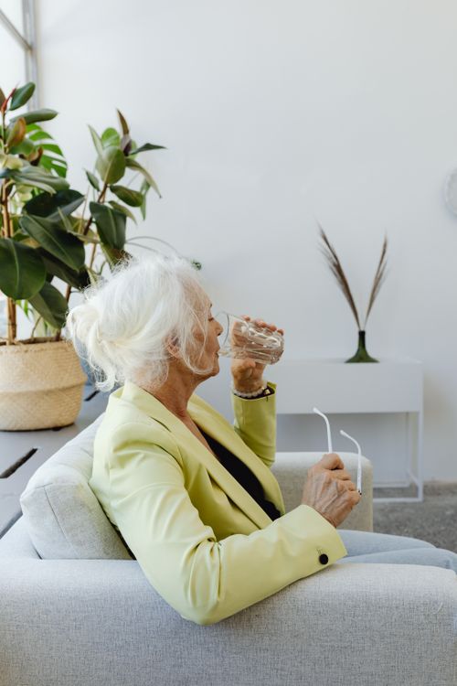 Free Elderly woman in bright blazer drinking water while sitting indoors. Stock Photo