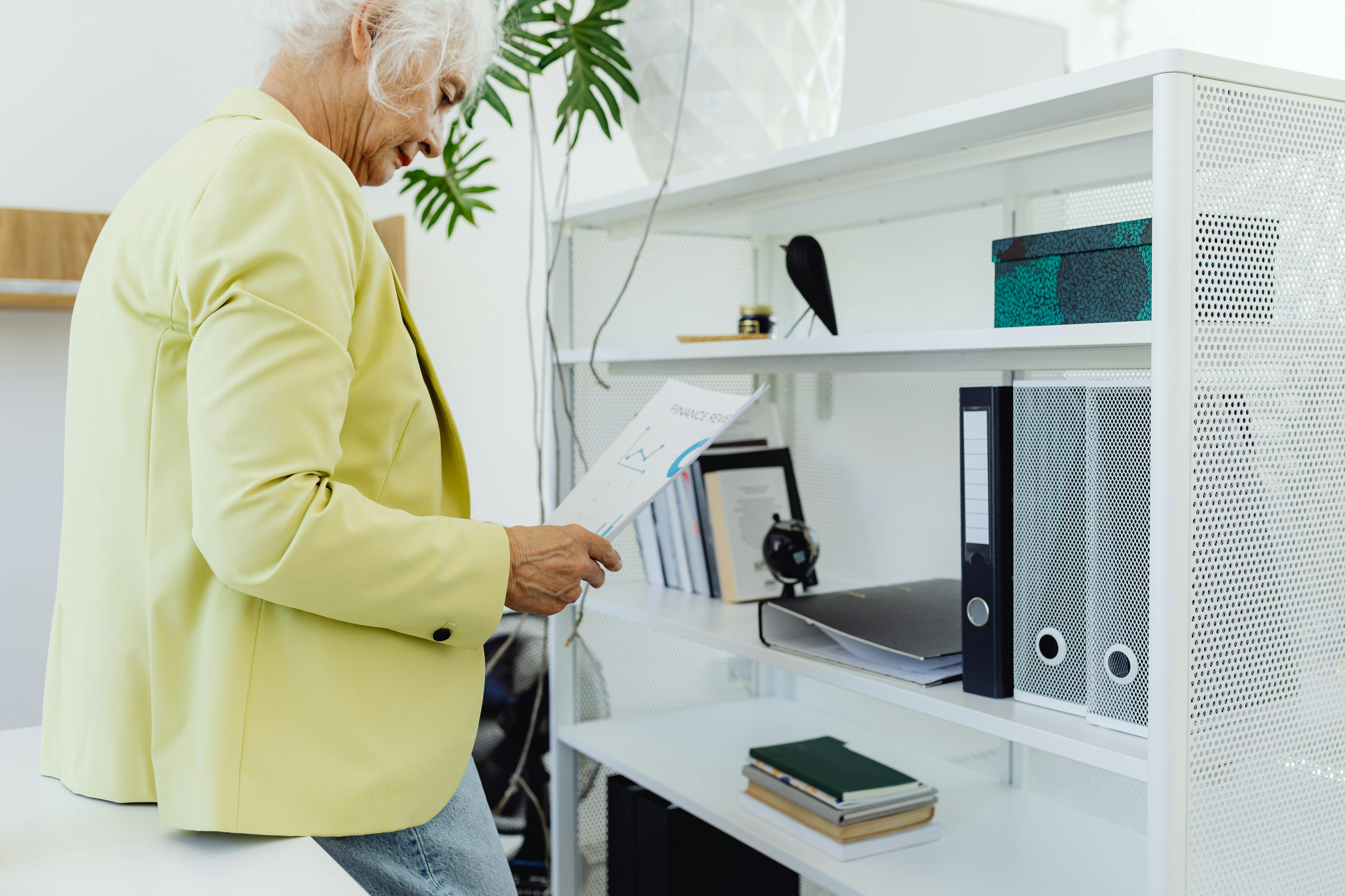 Elderly woman in yellow blazer reviews paperwork while leaning against a white bookcase in an office setting.