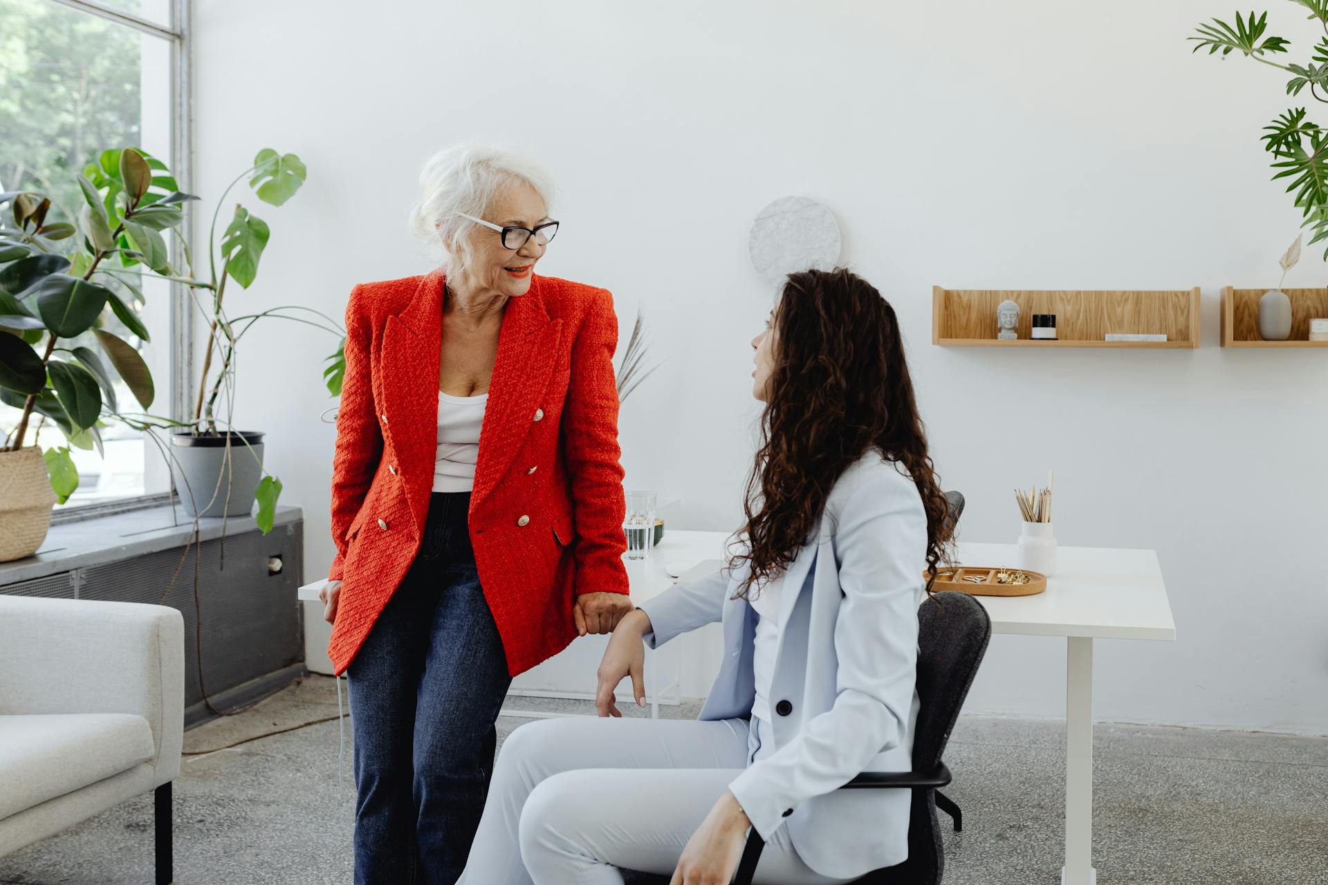 Two businesswomen in conversation in a stylish office space with plants and minimal decor.