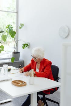 Elderly woman with white hair working at a desk in a bright, modern office environment.