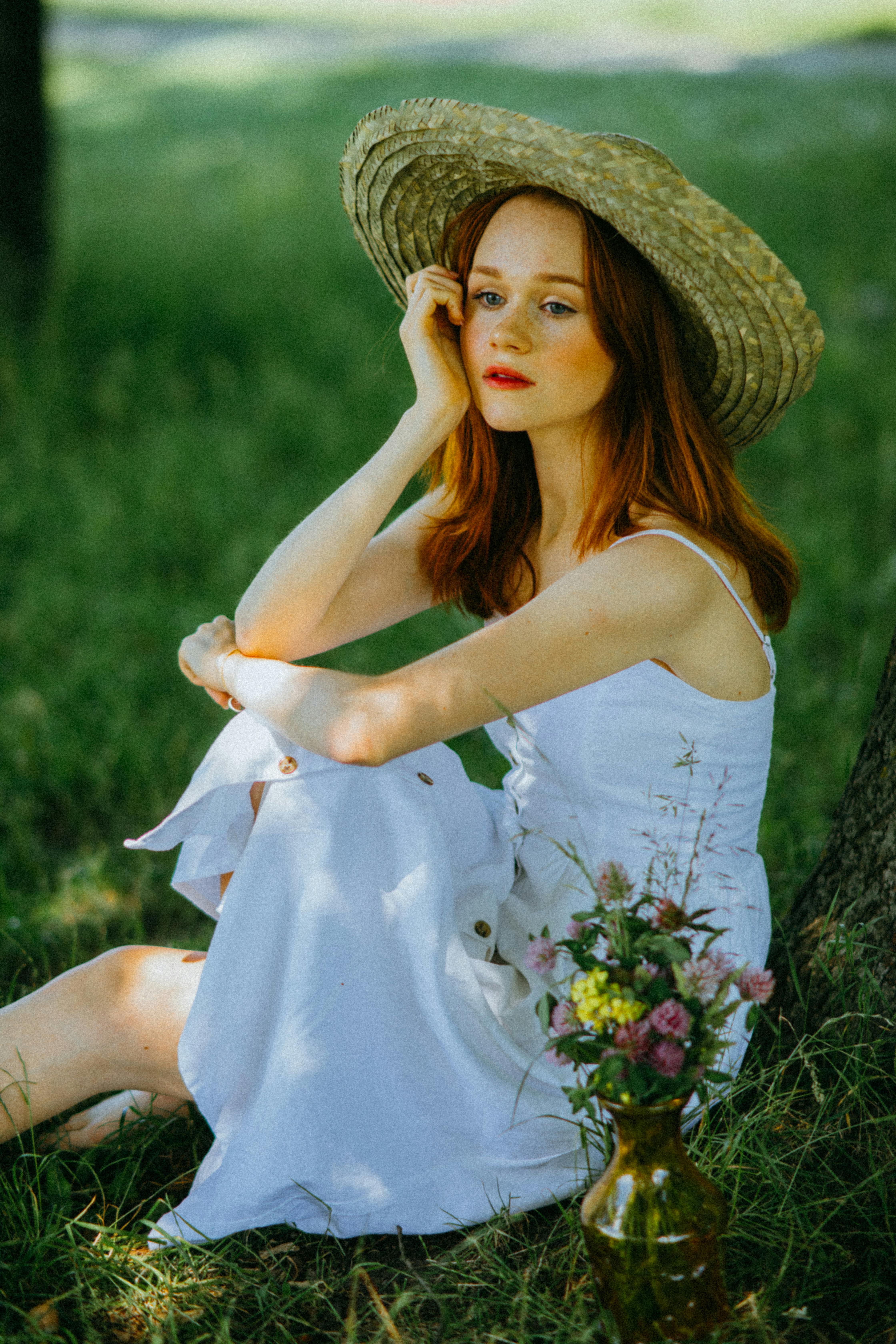 A woman in a white dress sits by a tree, wearing a straw hat, with a vase of flowers.