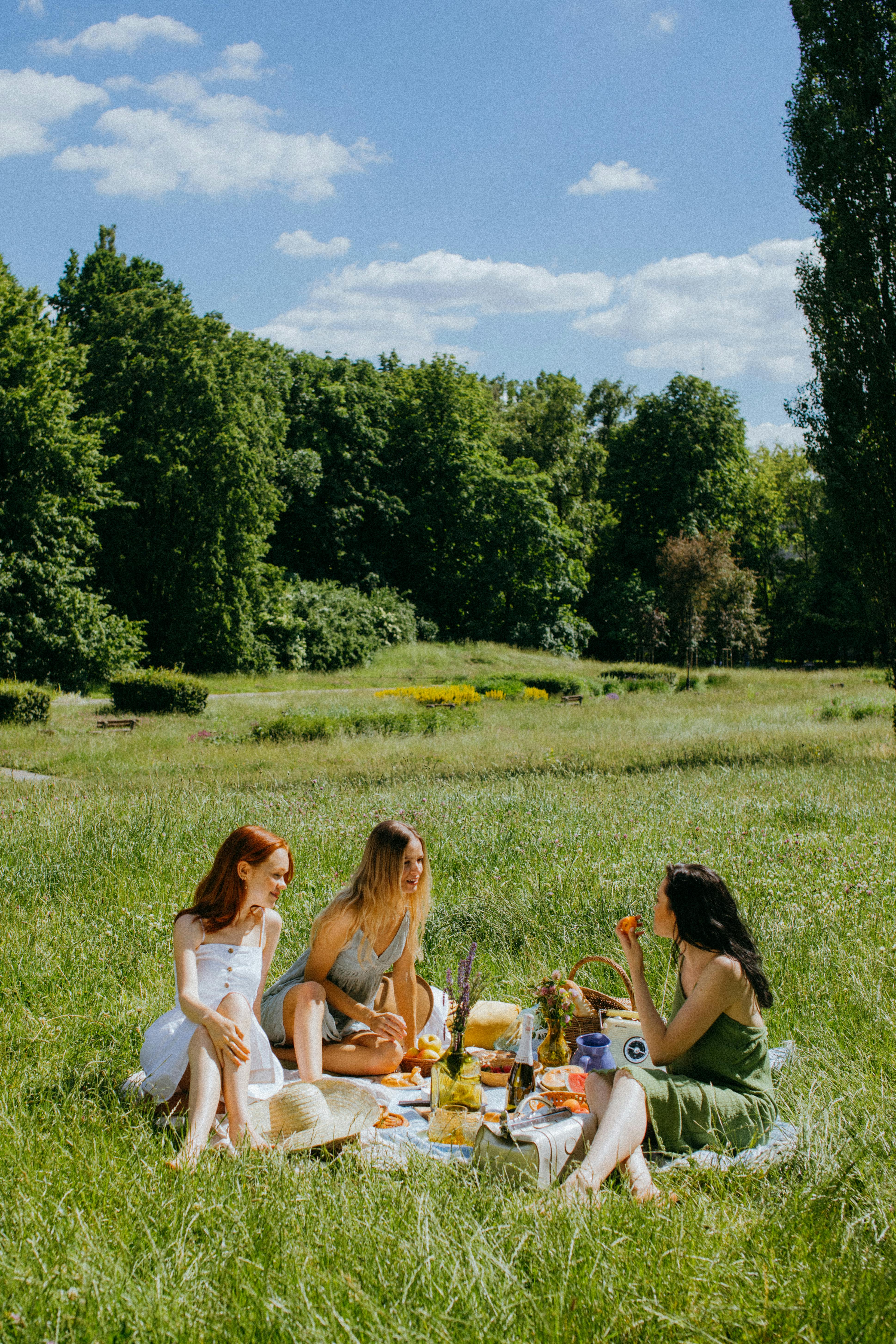 Three women enjoying a relaxed picnic in a sunny park, embracing nature and leisure.