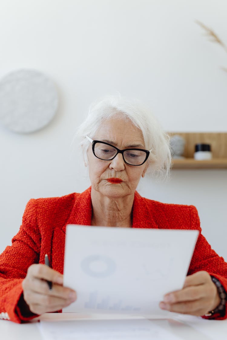 An Elderly Woman In Red Blazer Reading A Piece Of Document