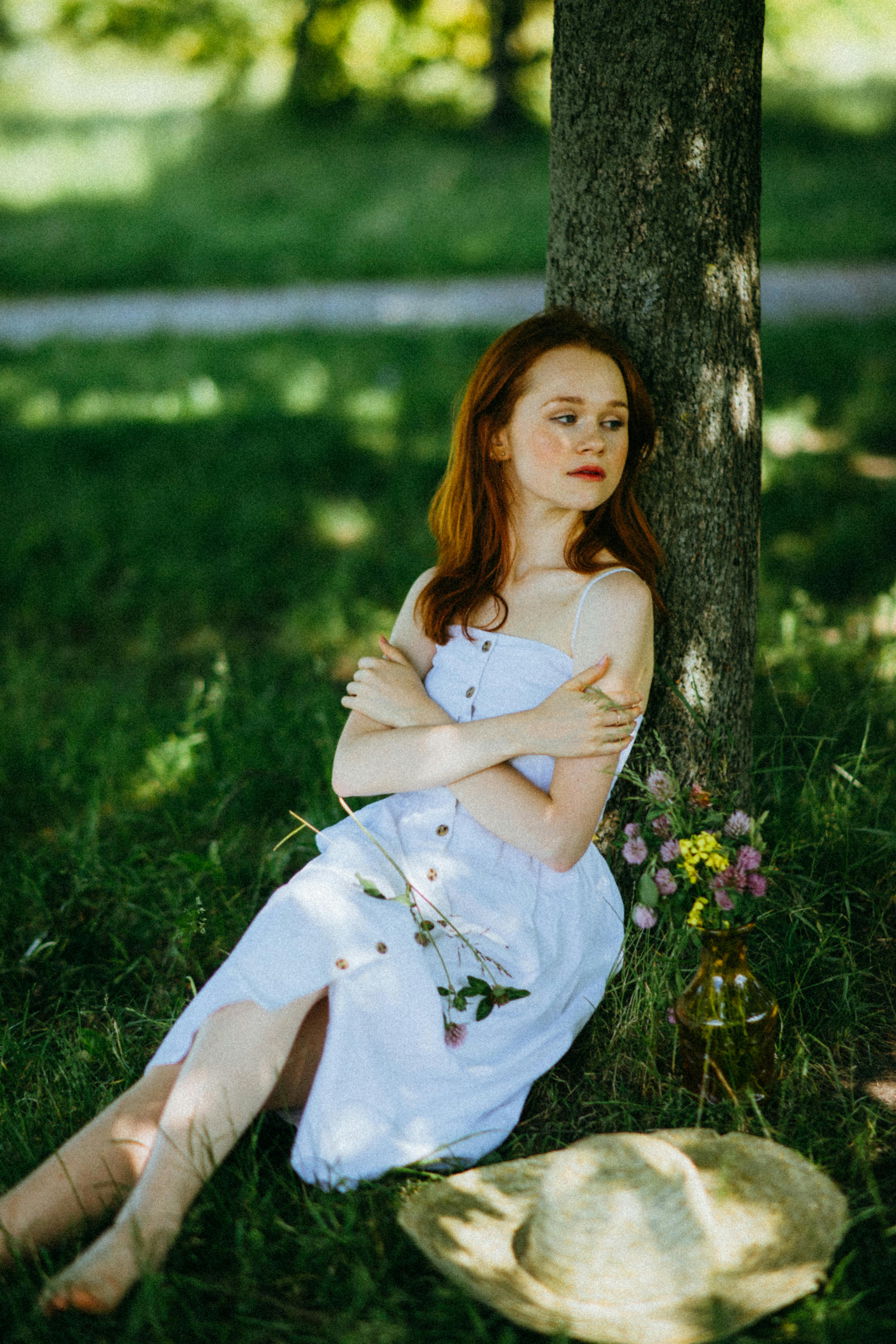 Woman in white dress sits under a tree, enjoying summer shade with a straw hat nearby.