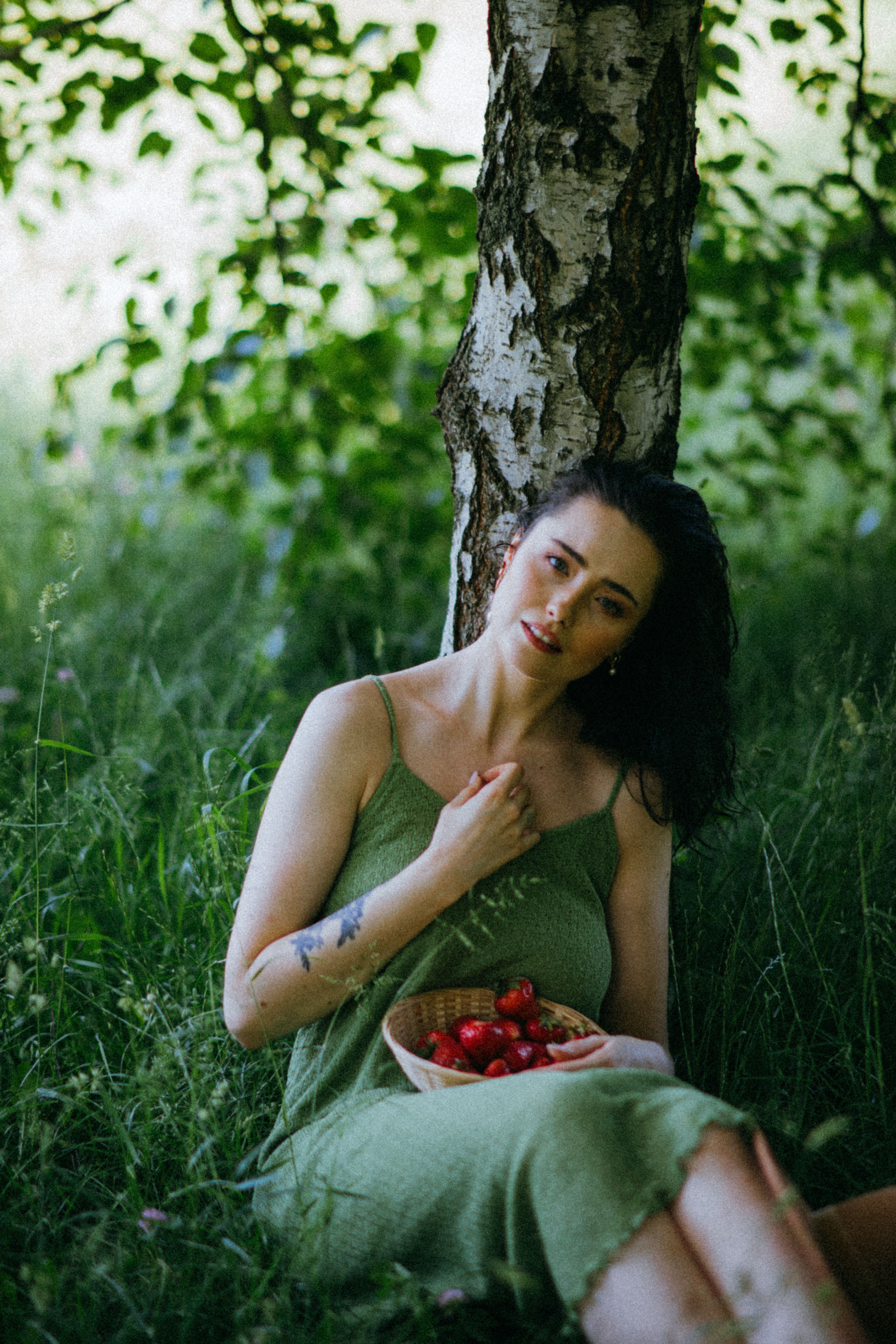 A woman in a green dress sits under a tree, holding a basket of strawberries, enjoying a peaceful summer day.