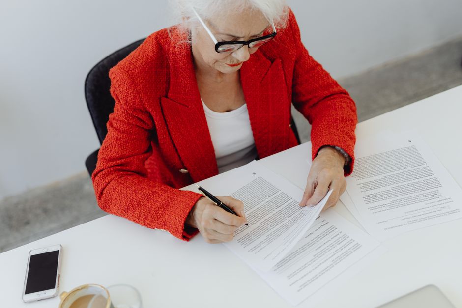 Mujer de negocios anciana con chaqueta roja firmando documentos en el escritorio, mostrando liderazgo.