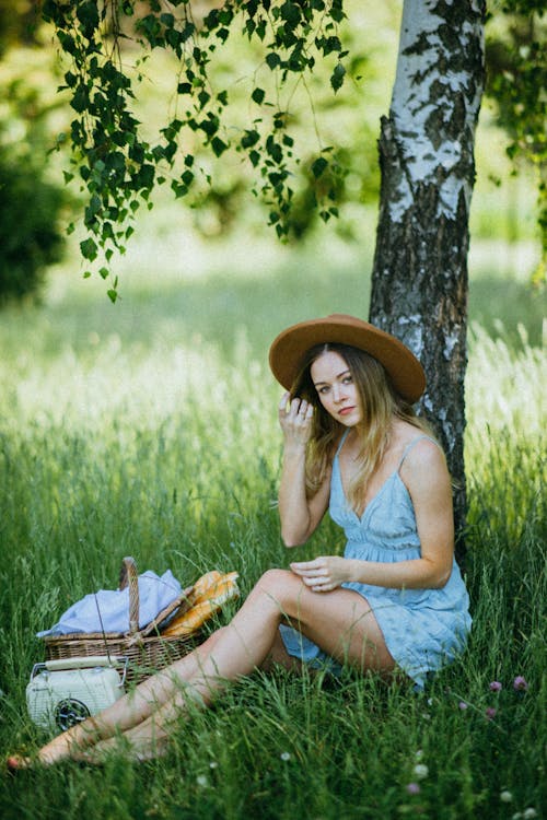 Woman in Blue Sleeveless Dress Wearing Brown Hat white Sitting on the Grass
