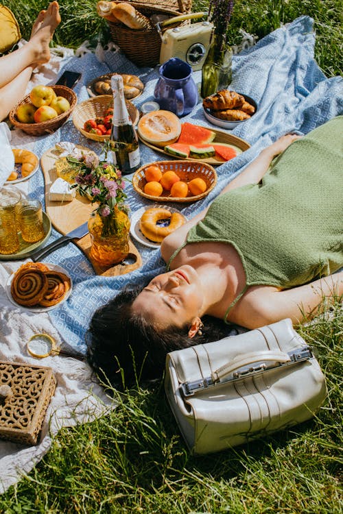 A Woman in Green Spaghetti Strap Dress Lying on Picnic Blanket with Food and a Bottle of Wine