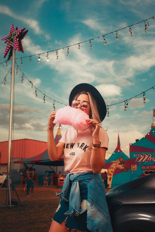 Blonde in a Hat Eating a Pink Cotton Candy, and Circus in Background