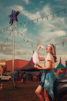 Smiling woman savoring candy floss at an outdoor carnival with vibrant tents.