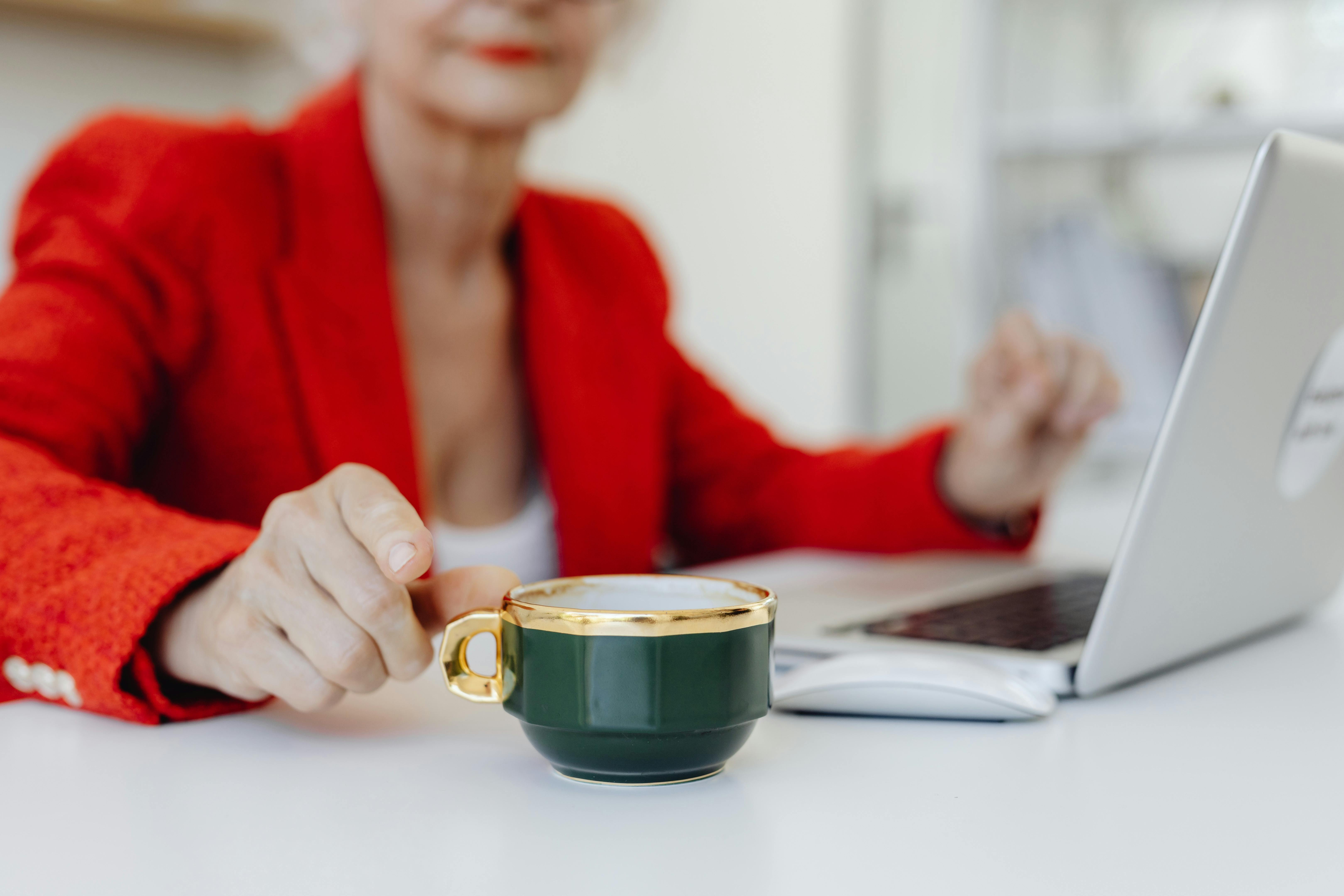 Close-up of a woman in a red blazer at a desk with coffee and laptop.