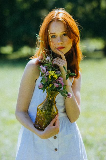 Young woman in a white dress holding a vase of wildflowers outdoors on a sunny day.