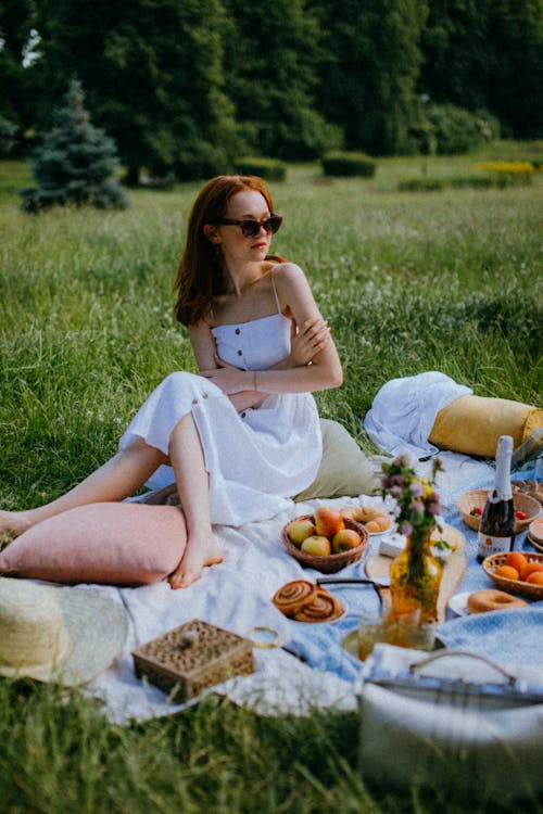 Woman in White Dress Sitting on a Picnic Blanket Surrounded  by Baskets of Snacks