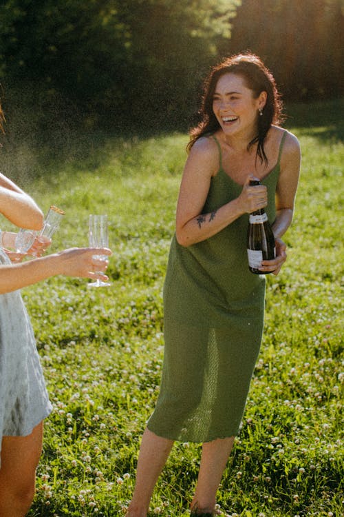 A Happy Woman in Green Dress Holding a Bottle of Wine while Standing on a Grass Field