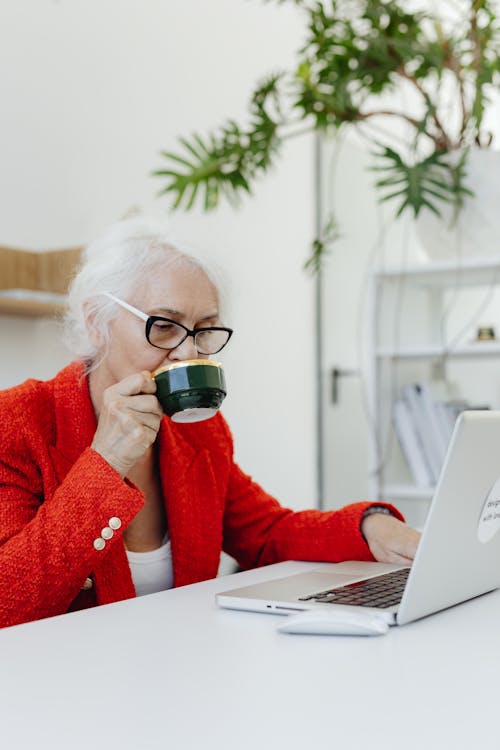 Free Woman in Red Coat Using Laptop while Drinking a Cup of Hot Beverage Stock Photo