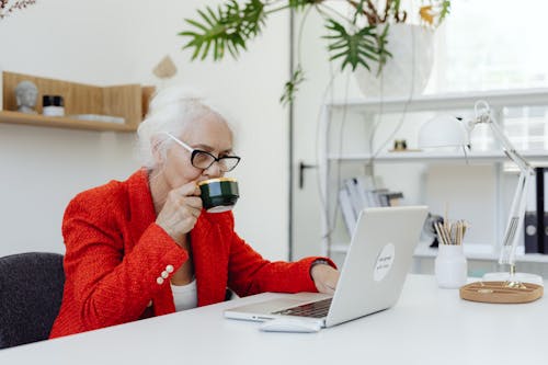 Free Woman in Red Coat Drinking Hot Beverage on a Cup while Looking at the Screen of a Laptop Stock Photo