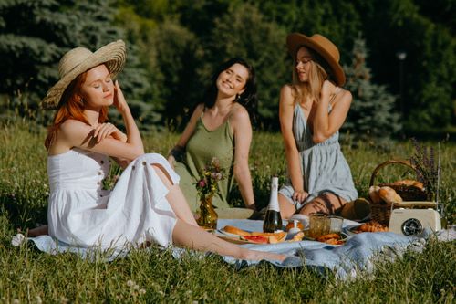 Free Three friends enjoying a sunny picnic outdoors with food, laughter, and nature. Stock Photo
