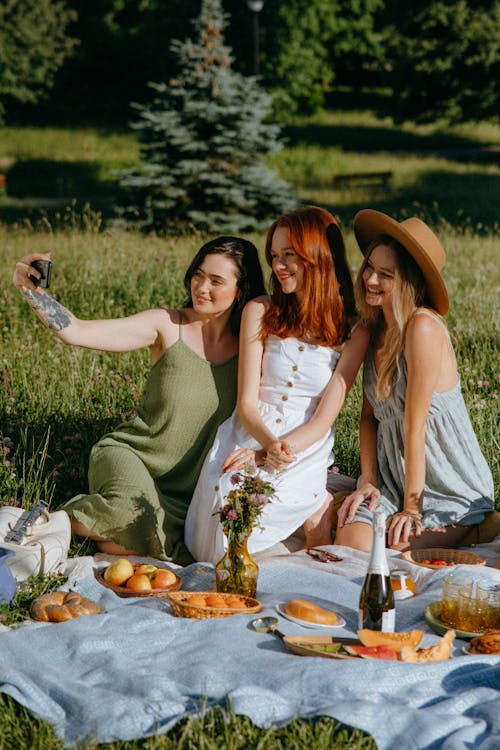 Free Women Sitting on a Picnic Blanket while Taking a Group Selfie Stock Photo