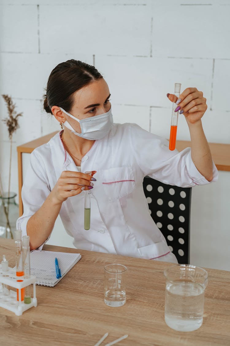 Woman In White Lab Gown Wearing Face Mask While Holding A Test Tubes
