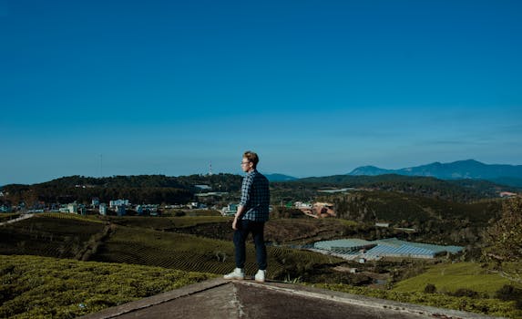 A man stands outdoors overlooking a picturesque countryside landscape under a clear blue sky.