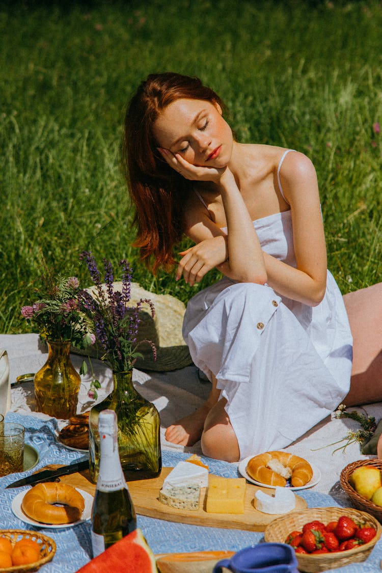 A Woman In White Dress Sitting On A Picnic Blanket Surrounded By Foods