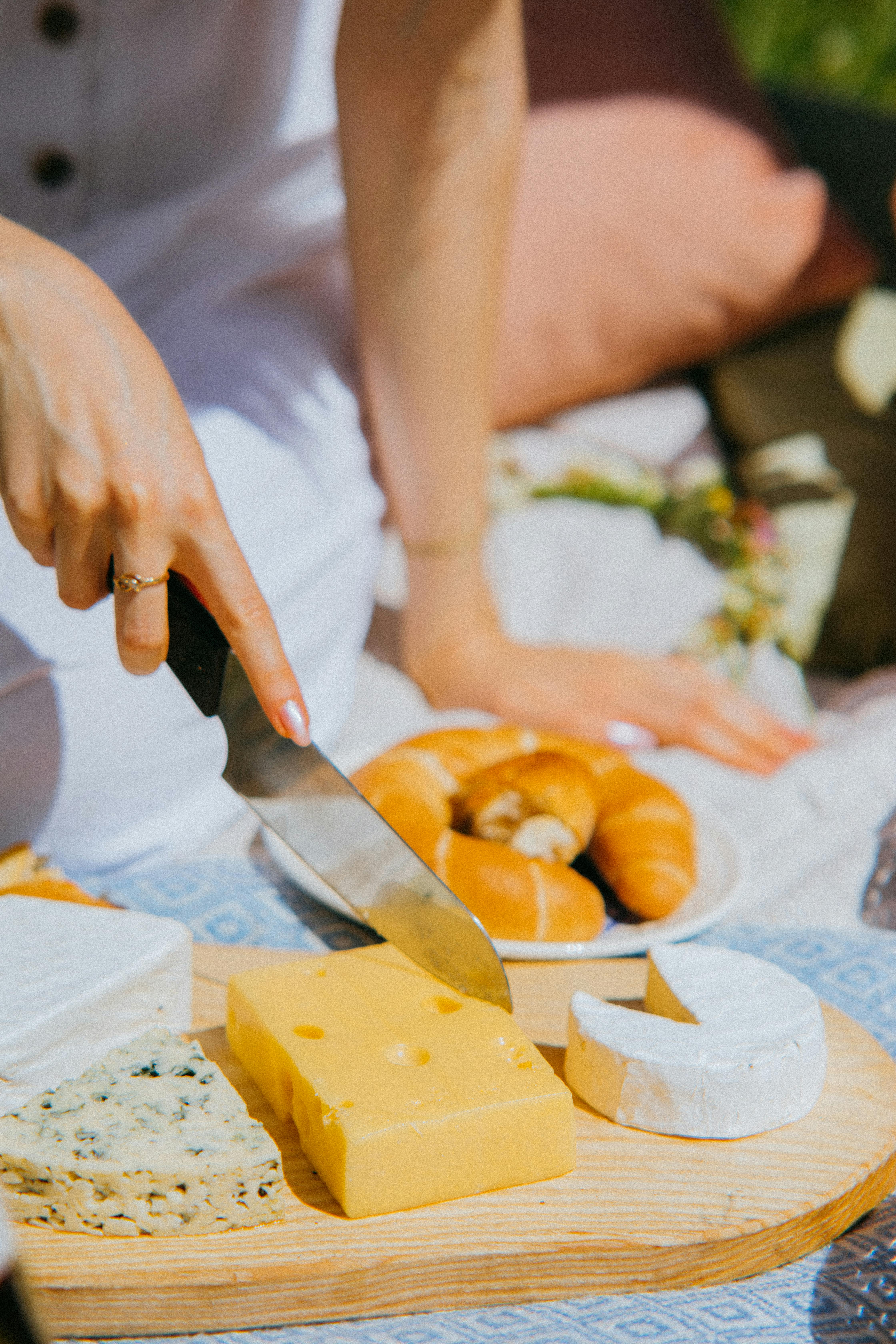 A close-up of a hand slicing a variety of cheeses outdoors. Perfect for lifestyle and food photography needs.