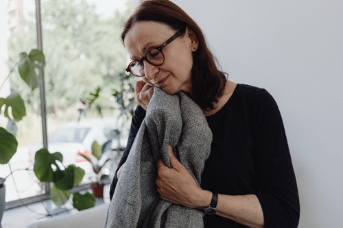 Free A senior woman wearing glasses stands indoors holding a gray blanket, deep in thought. Stock Photo