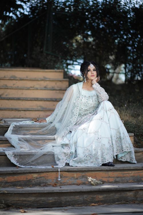 Free Beautiful bride in ornate white gown sitting on steps with hand on chin, serene outdoor setting. Stock Photo