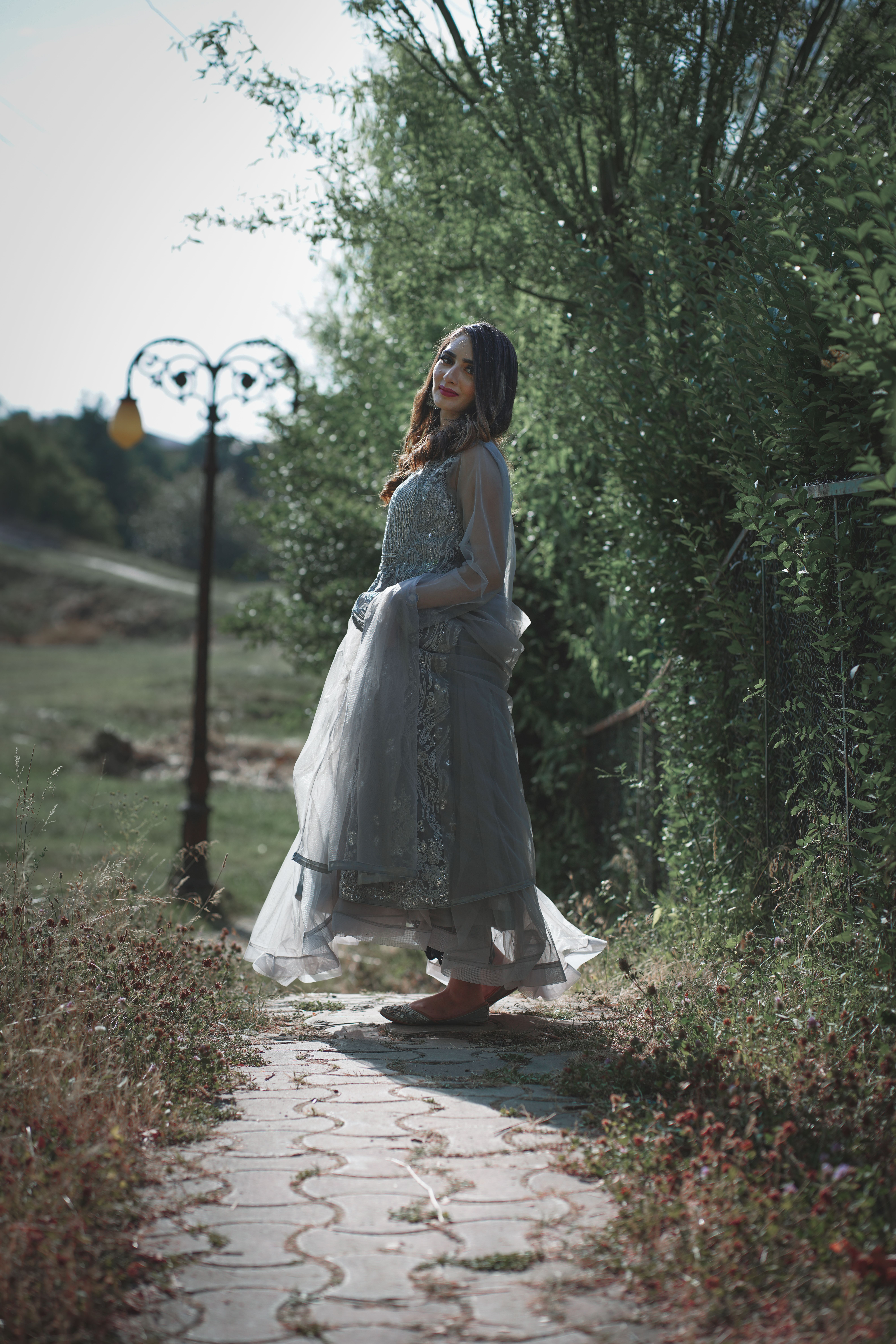 Elegant Indian woman poses in a scenic outdoor setting, wearing a traditional dress with natural greenery around.