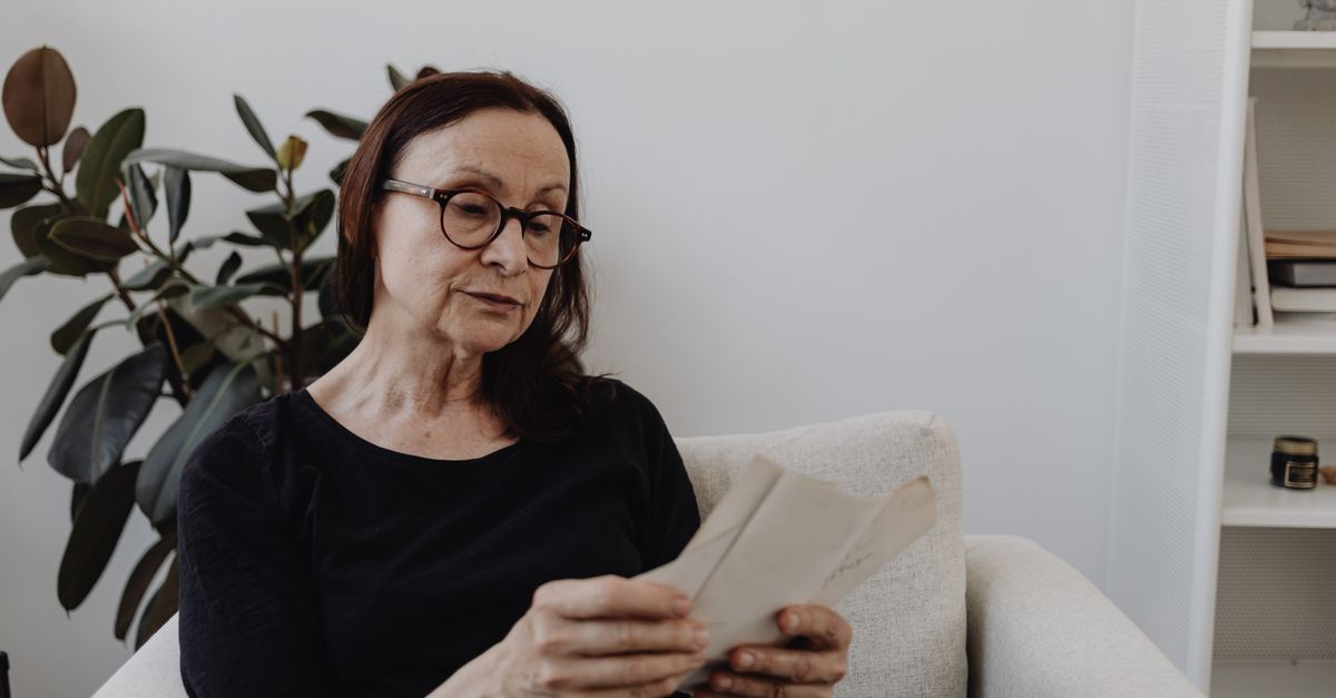Senior woman sitting in an armchair indoors, reading letters, reflecting on memories and emotions.