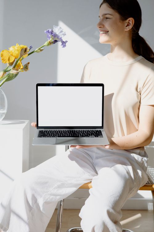 Free Smiling woman holding a blank screen laptop, sitting in a sunlit room with flowers. Stock Photo