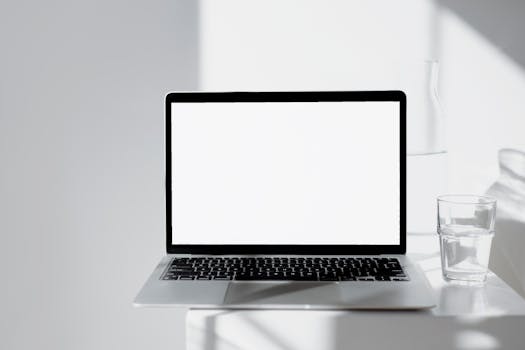 A sleek laptop on a white desk with a glass of water, bathed in natural light.
