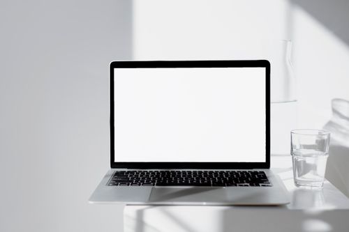 Free A sleek laptop on a white desk with a glass of water, bathed in natural light. Stock Photo