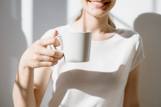 A smiling woman holds a white coffee cup in natural light indoors.