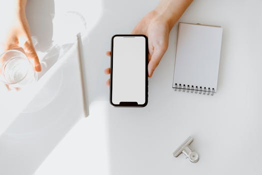 A clean, minimal workspace with a smartphone, glass of water, blank notepad, and pen on a white table.