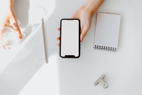 Free A clean, minimal workspace with a smartphone, glass of water, blank notepad, and pen on a white table. Stock Photo