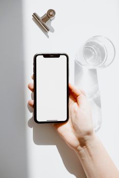 A hand holding a smartphone with a blank screen near a glass of water, perfect for technology and design mockups.
