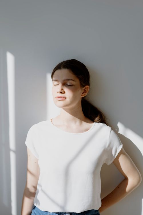 Free A serene portrait of a woman standing against a wall, eyes closed, in soft natural light. Stock Photo