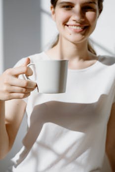 A cheerful woman holds a white mug, enjoying a moment indoors with natural sunlight.