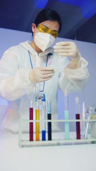Asian female scientist in PPE examines colorful liquids in lab.