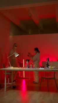 Two scientists in protective suits work in a red-lit laboratory, examining samples and conducting research.