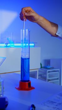 A scientist uses a stirring rod in a glass graduated cylinder filled with blue liquid.