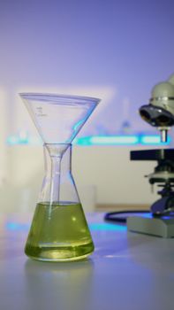 Close-up of an Erlenmeyer flask with green liquid and a microscope in a science lab.