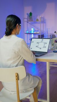 A scientist in a lab coat examines a chemical diagram on a laptop, surrounded by laboratory equipment.