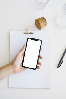A hand holds a smartphone with a blank screen above a clipboard on a clean, white desk.