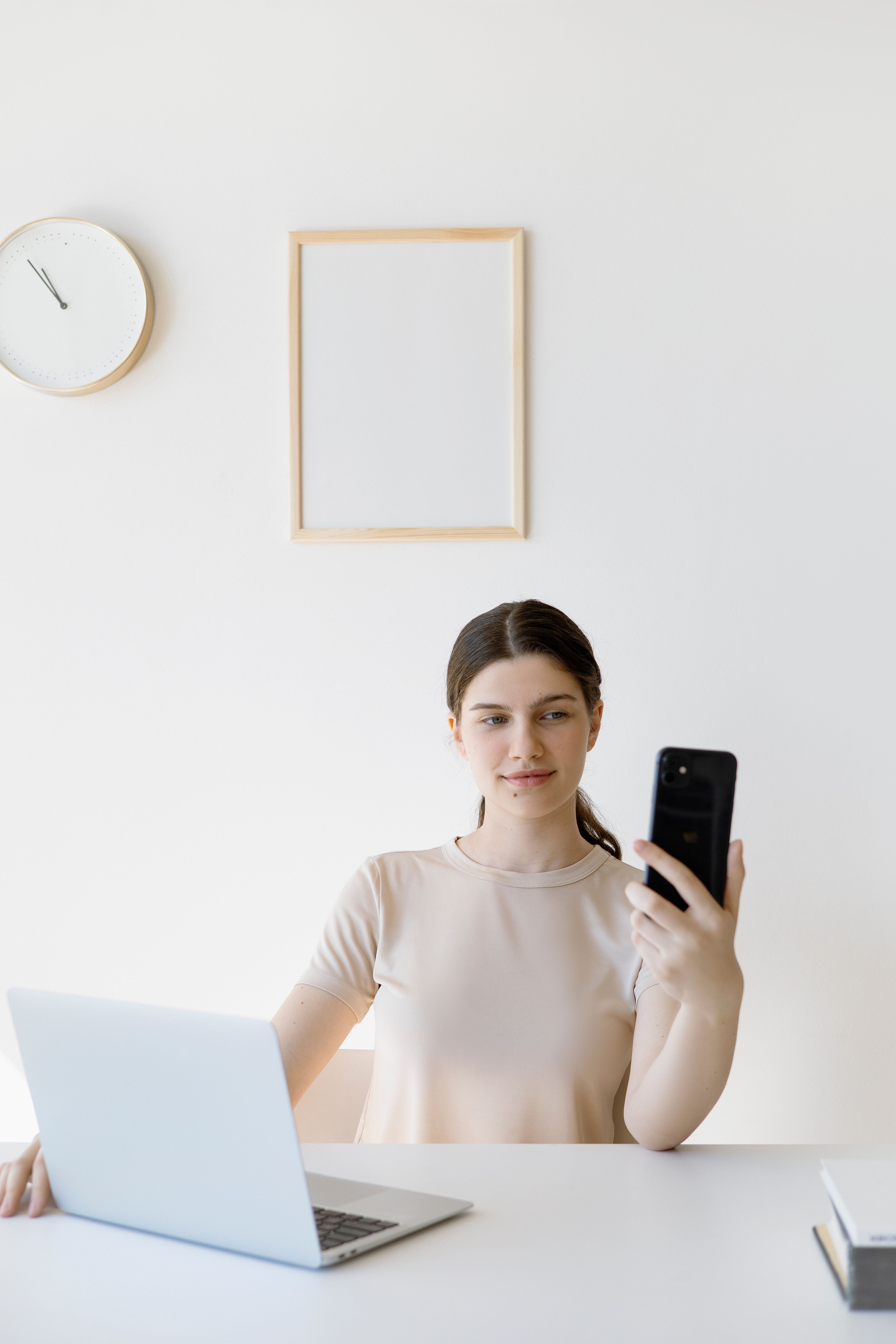 Young woman using a smartphone while working on a laptop at a minimalist desk.