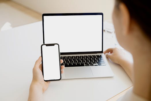 A woman holding a smartphone in front of a laptop, creating a seamless tech environment.