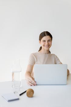 A woman smiling while working on her laptop at a desk with a minimalist setup.